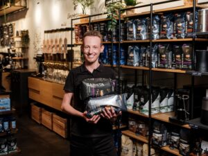 Lehrlingsportal.at Eine Person steht in einem Café und hält drei Beutel Barista-Kaffee in der Hand. Im Hintergrund sind Regale mit Kaffeeprodukten zu sehen.