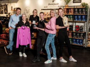 Lehrlingsportal.at Sechs Personen stehen in einem Café. Einige halten Produkte in den Händen, darunter ein rosa Hemd, eine Kaffeemühle und einen Kaffeebeutel. Im Hintergrund sind Regale mit Kaffeebeuteln und eine Theke zu sehen.