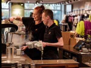 Lehrlingsportal.at Zwei Baristas in schwarzen Uniformen bereiten hinter einer Theke Kaffee zu. Einer dampft Milch auf, während der andere mehrere Tassen hält. Im Hintergrund ist ein Einzelhandelsgeschäft zu sehen.
