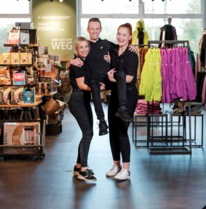 Lehrlingsportal.at Drei Personen in schwarzen Outfits posieren und lächeln in einem Einzelhandelsgeschäft. Zwei Erwachsene stehen mit einem Kind zwischen sich. Im Hintergrund sind Kleiderständer und Schaufenster zu sehen.
