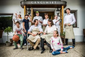 Lehrlingsportal.at Eine Gruppe von neun Personen, darunter Erwachsene und Kinder, posiert lächelnd vor einem Restauranteingang mit „Ganglwirt“-Schild, einige halten Küchenutensilien und ein Kind hält einen Basketball.