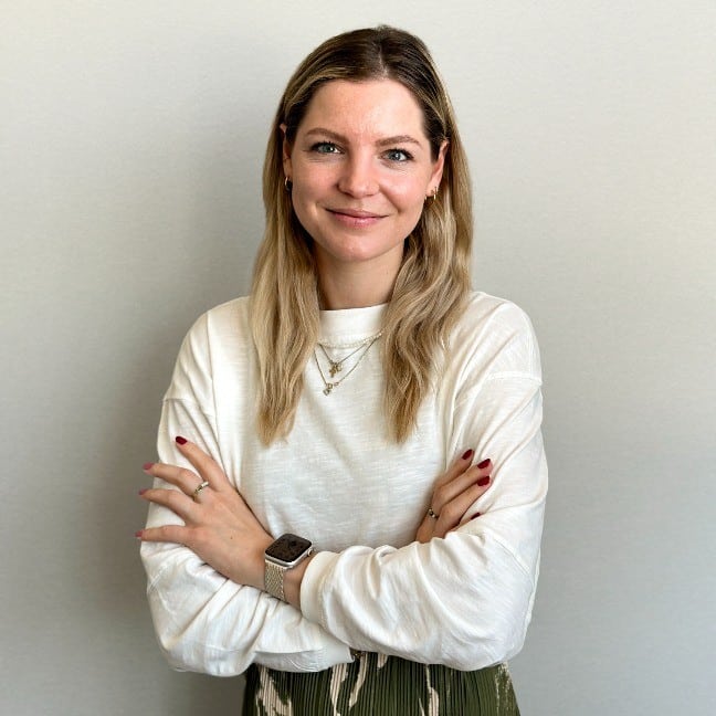 Lehrlingsportal.at Eine Frau mit langen blonden Haaren, einem weißen Langarmshirt und Schmuck bekleidet, steht mit verschränkten Armen vor einer schlichten hellen Wand und lächelt in die Kamera.