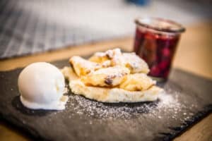 Lehrlingsportal.at Ein Dessert mit einer Kugel Vanilleeis, mit Puderzucker bestäubten Gebäckstücken und einem Gläschen rotem Grütze auf einem schwarzen Schieferteller.