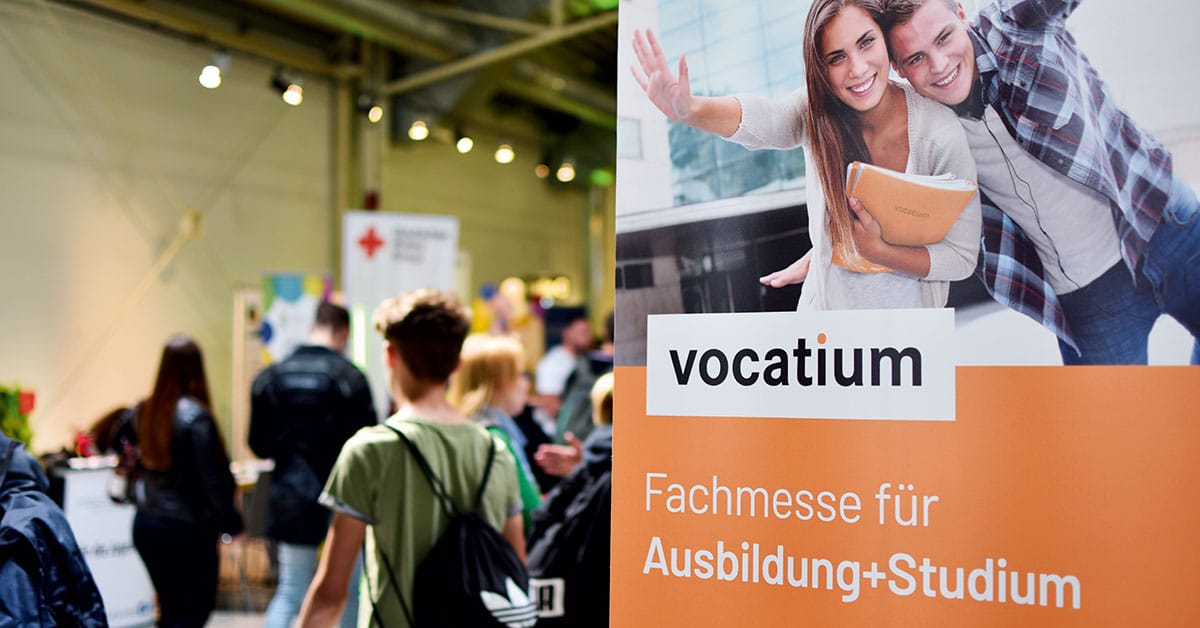 Lehrlingsportal.at Menschen besuchen eine Bildungsmesse. Im Vordergrund steht ein Schild mit der Aufschrift „vocatium Fachmesse für Ausbildung+Studium“ und einem Foto von zwei lächelnden Schülern.