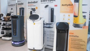 Lehrlingsportal.at Mehrere Restaurantservice-Roboter werden vor Werbebannern ausgestellt, auf denen jeweils unterschiedliche Modelle und Funktionen vorgestellt werden.