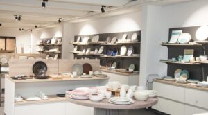 Lehrlingsportal.at In einem Ausstellungsraum sind verschiedene Keramikteller, -schalen und -tassen ordentlich auf Regalen und Tischen unter heller Beleuchtung angeordnet.
