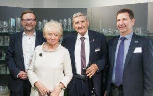 Lehrlingsportal.at Vier Erwachsene, drei Männer und eine Frau, stehen gemeinsam in einem Innenraum vor Regalen mit Glaswaren, sind formell gekleidet und lächeln in die Kamera.