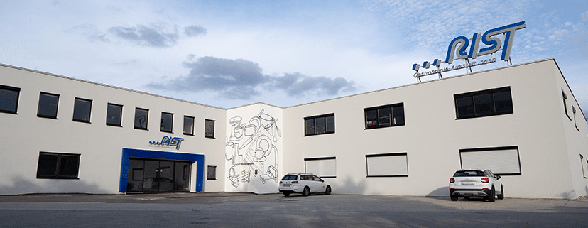 Lehrlingsportal.at Ein weißes Geschäftsgebäude mit dem Logo "AIST" an der Fassade, zwei geparkte Autos und ein Wandgemälde mit Küchenutensilien an einer Außenwand.
