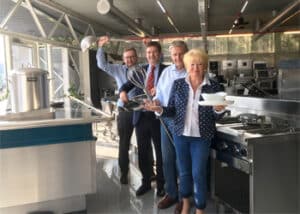 Lehrlingsportal.at Vier Personen stehen in einer Großküche, lächeln und halten verschiedene Küchenutensilien und Gerichte in der Hand, im Hintergrund sind Geräte aus Edelstahl zu sehen.