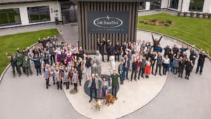 Lehrlingsportal.at Eine große Gruppe von Menschen steht vor dem Gut Brandlhof Hotel & Conference und posiert und winkt für ein Gruppenfoto neben einem Schild und zwei Hunden.