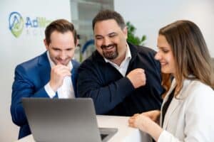 Lehrlingsportal.at Drei professionell gekleidete Personen stehen an einem Schreibtisch, lächeln und schauen auf einen Laptop-Bildschirm in einer modernen Büroumgebung.