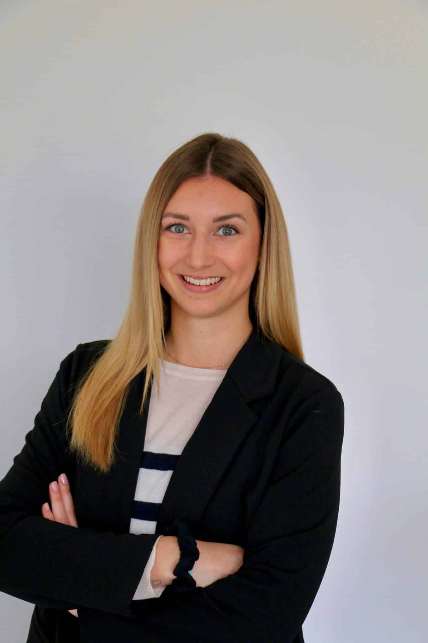 Lehrlingsportal.at Eine Frau mit glattem blondem Haar, die einen schwarzen Blazer und ein gestreiftes Hemd trägt, steht mit verschränkten Armen und einem leichten Lächeln vor einem schlichten weißen Hintergrund.
