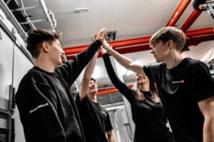 Lehrlingsportal.at Vier junge Erwachsene in schwarzen Hemden geben sich in einer Industriehalle mit roten Rohren über dem Kopf gegenseitig High Fives.