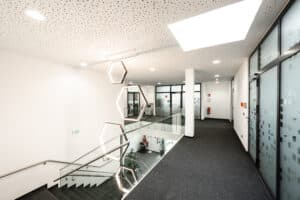 Lehrlingsportal.at Moderner Flur mit Glaswänden, sechseckigen Beleuchtungskörpern und einer Treppe, die nach unten führt; der Raum ist mit Teppichboden und weißen Decken mit perforierten Paneelen ausgestattet.