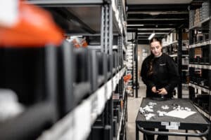 Lehrlingsportal.at Eine Frau steht an einem Wagen und sortiert kleine weiße Plastikteile in einem Lagergang, der mit beschrifteten Behältern und Regalen gesäumt ist.
