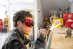 Lehrlingsportal.at Eine Person, die eine Schutzbrille trägt, schweißt in einer Werkstatt mit einem Schweißbrenner an einem Metallstück, wobei Funken und Rauch sichtbar sind.