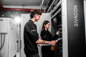 Lehrlingsportal.at Zwei Techniker arbeiten in einem Serverraum mit der Aufschrift "SIVACON" an elektrischen Geräten. Der eine hält Dokumente in der Hand, der andere justiert Komponenten in einem Schrank.
