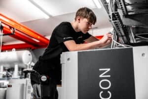 Lehrlingsportal.at Ein junger Techniker in Arbeitskleidung justiert Kabel an elektronischen Geräten in einer hell erleuchteten Industrieumgebung.