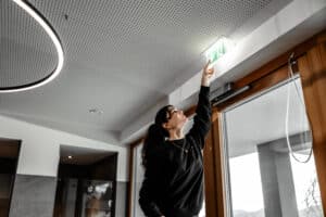 Lehrlingsportal.at Eine Frau steht auf Zehenspitzen und zeigt auf ein grün leuchtendes Notausgangsschild, das an der Decke über einer Tür angebracht ist.
