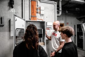 Lehrlingsportal.at Drei Personen stehen vor einer offenen elektrischen Schalttafel in einer Industrieumgebung und besprechen deren Bestandteile. Über der Schalttafel an der Wand sind orangefarbene Kabel zu sehen.