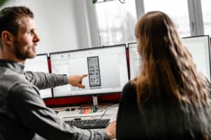 Lehrlingsportal.at Ein Mann zeigt auf einen Computermonitor, der technische Diagramme anzeigt, während eine Frau neben ihm an einem Arbeitsplatz mit mehreren Bildschirmen und Büromaterialien sitzt.