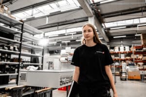 Lehrlingsportal.at Eine Frau in einem schwarzen T-Shirt geht durch ein Industrielager mit Regalen voller Kisten und Geräte im Hintergrund.