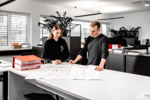 Lehrlingsportal.at Zwei Personen stehen an einem Schreibtisch in einem modernen Büro und betrachten die vor ihnen ausgebreiteten Baupläne und Dokumente.