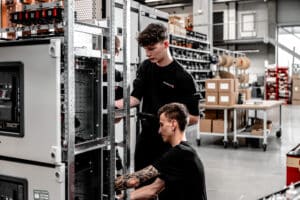 Lehrlingsportal.at Zwei Personen arbeiten gemeinsam an einem offenen Schaltschrank in einer mit Geräten, Regalen und Kartons gefüllten Industriehalle.