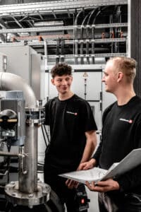 Lehrlingsportal.at Zwei Techniker in schwarzen Hemden stehen neben Industrieanlagen; einer hält ein Klemmbrett in der Hand, während sie in einer modernen Anlage Maschinen besprechen und überprüfen.