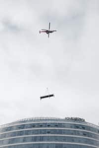 Lehrlingsportal.at Ein Hubschrauber hebt eine große Plattform über ein modernes Glasgebäude, auf dessen Dach das "Four Points"-Schild zu sehen ist, vor einem bewölkten Himmel.