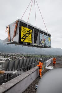 Lehrlingsportal.at Ein großes HLK-Gerät wird mit einem Kran auf ein Dach gehoben, während zwei Arbeiter in orangefarbener Schutzkleidung den Vorgang überwachen.