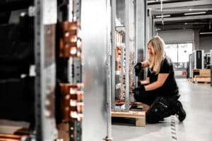 Lehrlingsportal.at Eine Frau mit Arbeitshandschuhen kniet auf dem Boden und prüft elektrische Geräte in einer Industrieumgebung.