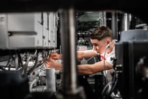 Lehrlingsportal.at Ein junger Mann arbeitet an Maschinen und benutzt Werkzeuge, um Geräte in einer industriellen Umgebung einzustellen oder zu reparieren.