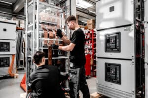 Lehrlingsportal.at Zwei Techniker in schwarzer Arbeitskleidung montieren oder reparieren elektrische Geräte in einer Industrieanlage und verwenden dabei Werkzeuge an einem großen Metallgestell mit Kabeln und Bauteilen.