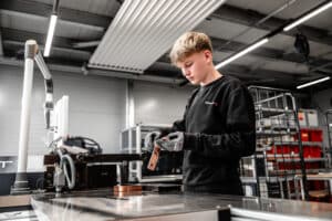 Lehrlingsportal.at Ein junger Mensch mit Handschuhen und schwarzem Sweatshirt arbeitet in einer industriellen Werkstatt mit einem Metallteil.