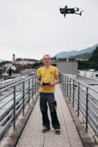 Lehrlingsportal.at Ein Mann in einem gelben Hemd steuert eine Drohne auf einer Dachterrasse, während im Hintergrund Berge und Gebäude zu sehen sind.