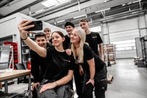 Lehrlingsportal.at Sechs junge Leute in schwarzen Hemden posieren für ein gemeinsames Selfie in einer geräumigen Industriehalle, in der im Hintergrund Regale und Geräte zu sehen sind.