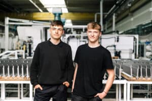 Lehrlingsportal.at Zwei junge Männer in schwarzen Hemden stehen Seite an Seite, der Kamera zugewandt, in einer Industrie- oder Fertigungsanlage mit Maschinen im Hintergrund.