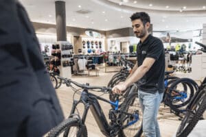 Lehrlingsportal.at Ein Mann in einem schwarzen Poloshirt steht neben einem Mountainbike in einem Fahrradgeschäft, umgeben von anderen Fahrrädern und Auslagen für Fahrradzubehör.