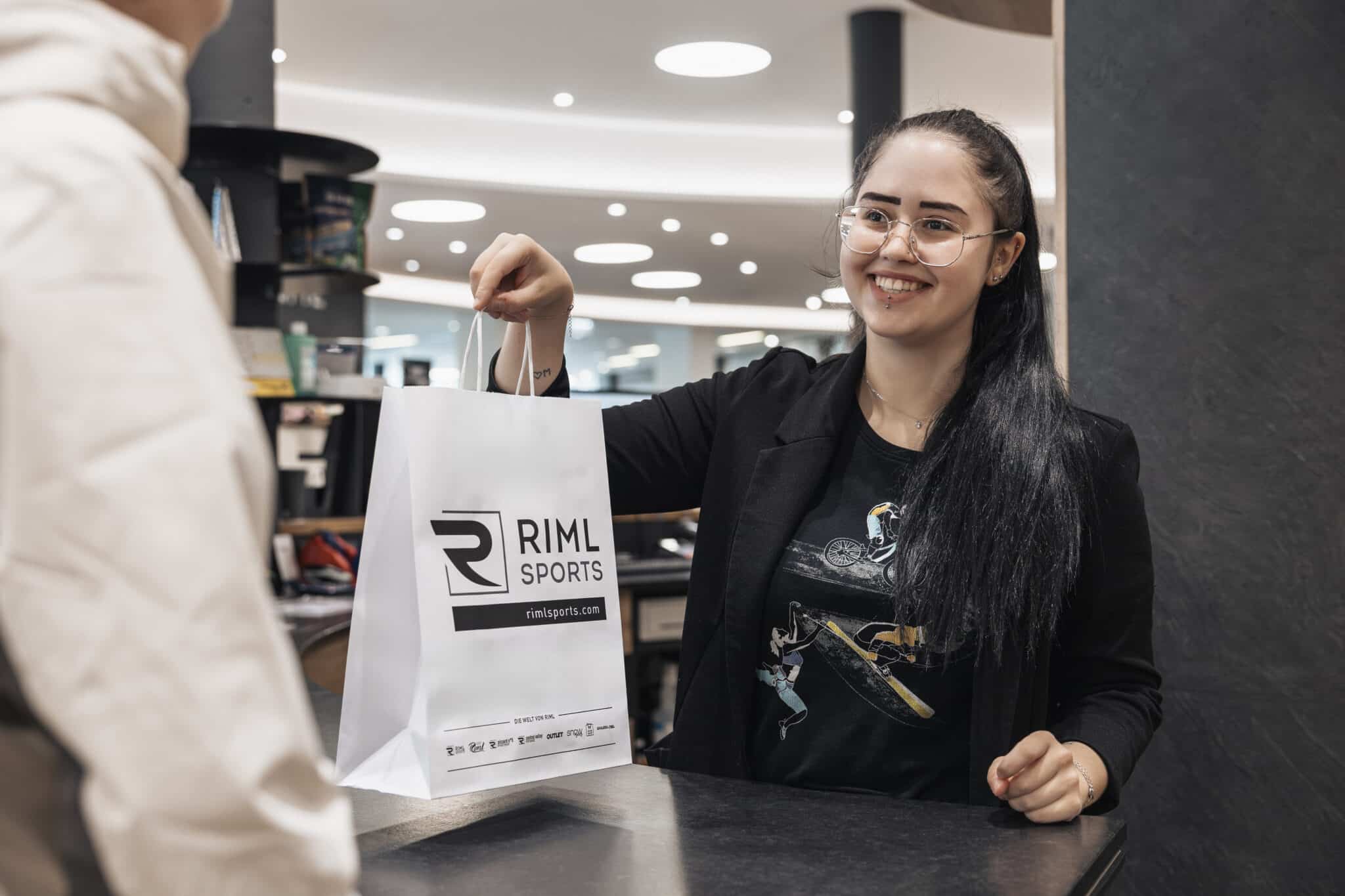 Lehrlingsportal.at Eine lächelnde Frau reicht einer Kundin in einem gut beleuchteten Geschäft eine Einkaufstasche von RIML Sports über den Tresen.