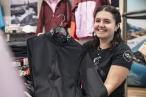 Lehrlingsportal.at Eine lächelnde Frau hält eine schwarze Jacke mit Reißverschluss auf einem Kleiderbügel in einem Bekleidungsgeschäft hoch.