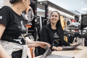 Lehrlingsportal.at Zwei Frauen stehen in einem Bekleidungsgeschäft an einer Theke und falten Kleidung, wobei sie einander anlächeln. Im Hintergrund sind Kleiderstangen und Regale zu sehen.