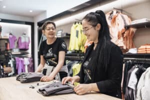 Lehrlingsportal.at Zwei Frauen, beide mit Brillen und dunklen Hemden, falten Kleidung auf einem Holztisch in einem hell erleuchteten Bekleidungsgeschäft mit Regalen voller bunter Kleidung im Hintergrund.