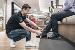 Lehrlingsportal.at Ein Mann kniet nieder, um einer sitzenden Person in einem modernen Einzelhandelsgeschäft beim Anpassen und Anziehen von Outdoor-Stiefeln zu helfen.