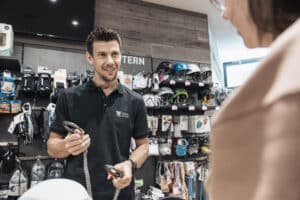 Lehrlingsportal.at Ein Angestellter in einem schwarzen Poloshirt zeigt einem Kunden in einem Geschäft für Outdoor-Ausrüstung zwei Kletterkarabiner.