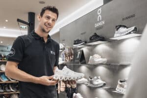 Lehrlingsportal.at Ein Mann in einem schwarzen Polohemd hält einen weißen Turnschuh in einem Schuhgeschäft in der Nähe einer Auslage mit verschiedenen Lauf- und Freizeitschuhen mit der Aufschrift "DAMEN".