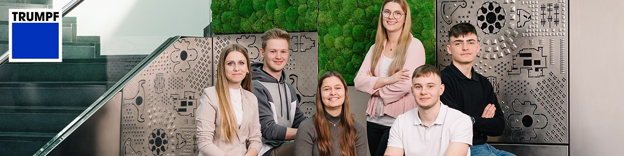 Lehrlingsportal.at Sechs junge Erwachsene posieren zusammen in einem Innenraum in der Nähe einer Treppe. Im Hintergrund sind eine grüne Wand, dekorative Metallplatten und ein TRUMPF Logo zu sehen.