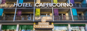 Lehrlingsportal.at Die vordere Fassade des Hotels Capricorno mit großen Schildern, bunten Tafeln an den Balkonen und dem von der Straße aus sichtbaren Eingang.