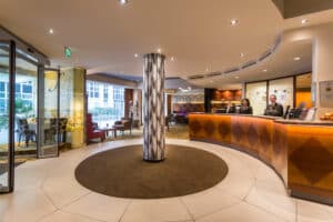 Lehrlingsportal.at Hotellobby mit zentraler Säule, geschwungener Holzrezeption, zwei Rezeptionisten, Sitzbereich und modernem Dekor.