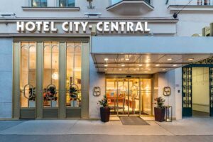 Lehrlingsportal.at Außenansicht des Hotels City Central mit großen gläsernen Eingangstüren und Fenstern mit warmer Beleuchtung, darunter ein Schild mit dem Namen des Hotels.